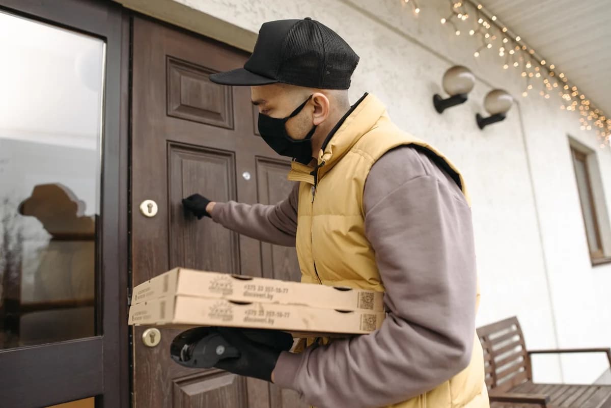 Deliveryman with mask and gloves knocking on door holding pizza boxes