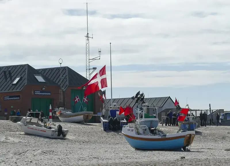 Danish flag flying on a pole near boats and buildings at a beach
