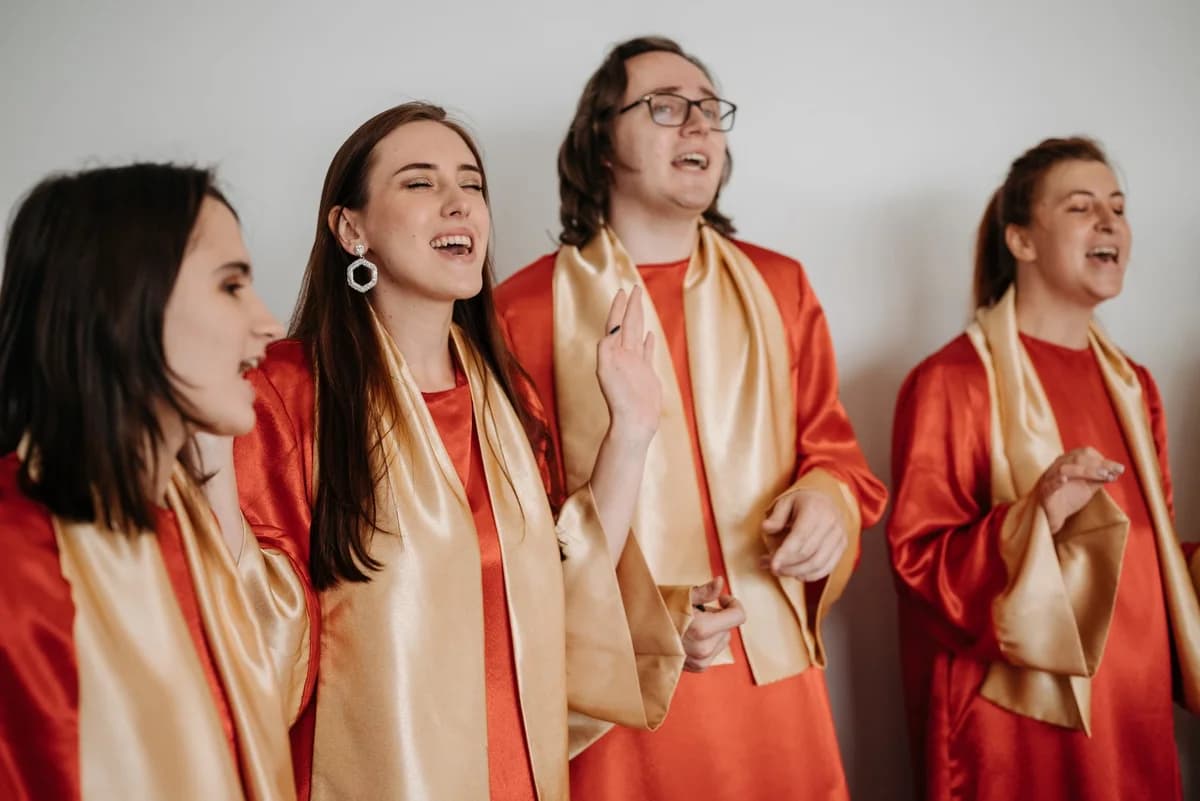 Four adults singing in red choir robes with gold scarves indoors.