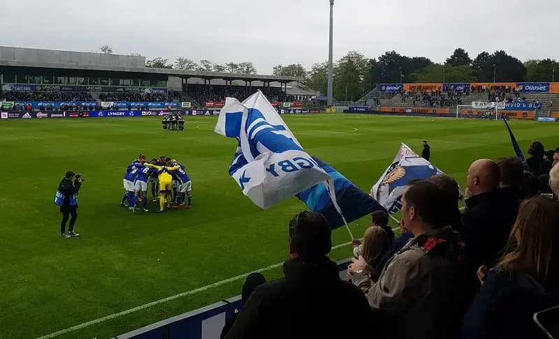 Football teams huddling on the field with fans and flags in the foreground