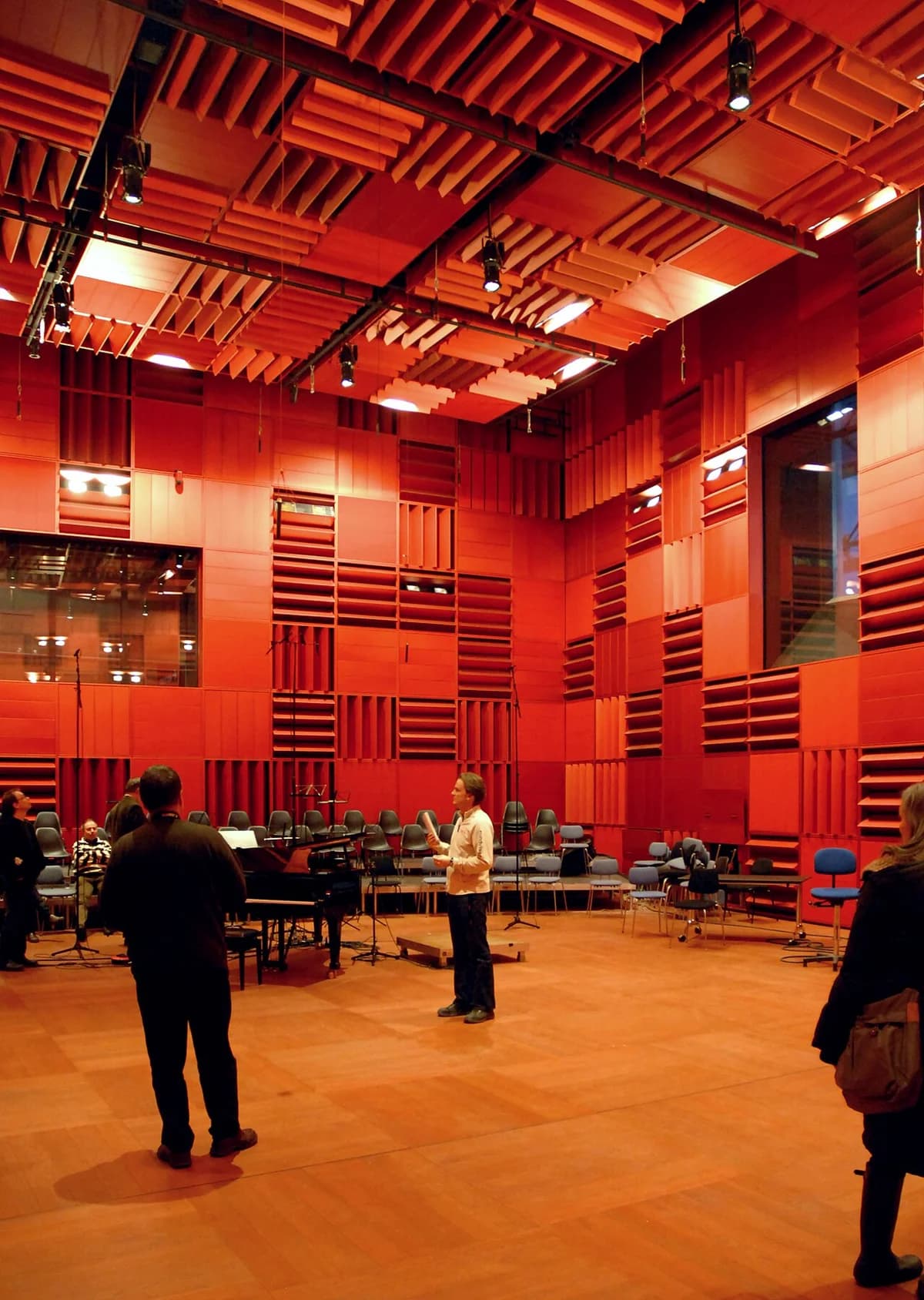 Interior of a red acoustic concert hall with people standing around and a piano.