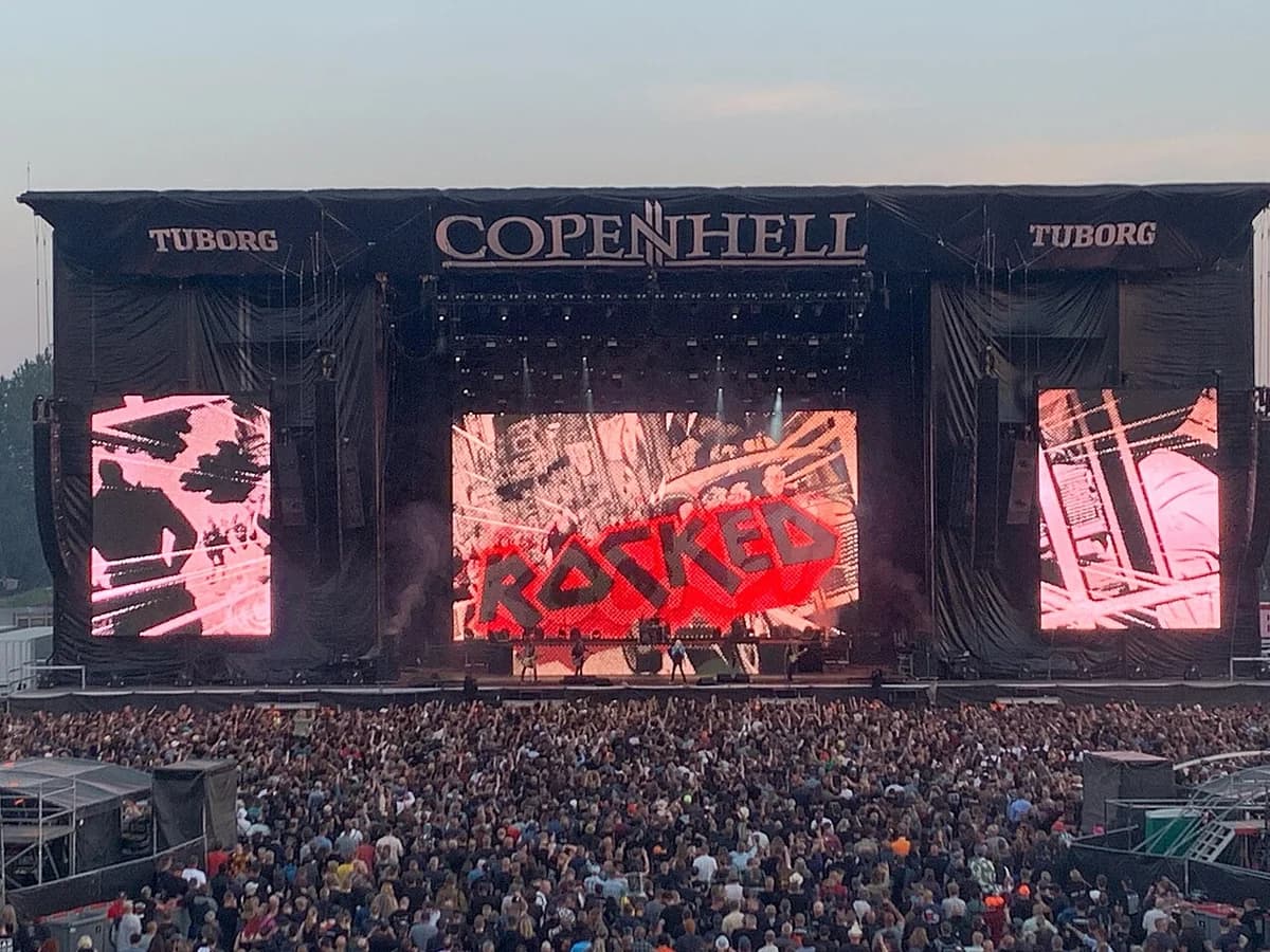A crowd watches a rock festival stage at Copenhell with "ROCKED" on the main screen.
